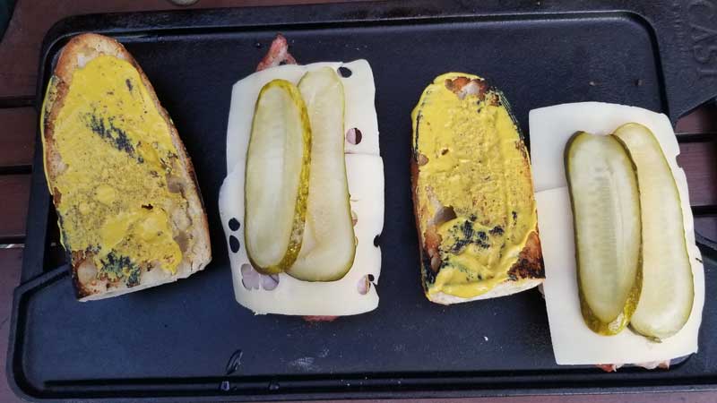 Baguette top covered in mustard and bottom covered with meat, cheese, and pickles.